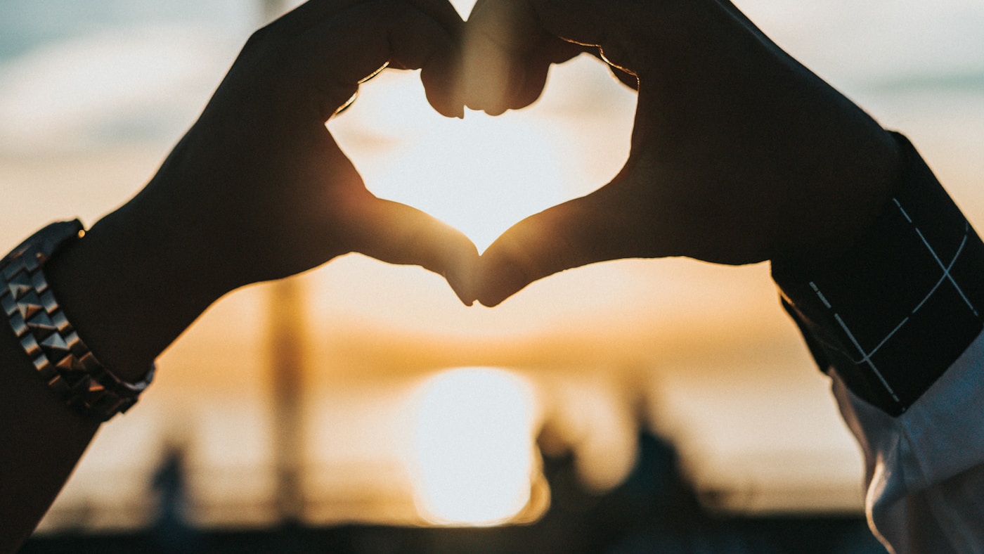 Two people forming a heart shape with their hands against a warm sunset sky